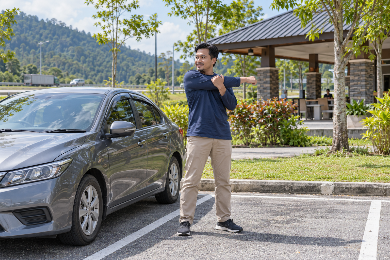 Pemandu lelaki berpakaian sopan melakukan regangan bahu ringan di sebelah kereta yang diparkir di kawasan rehat pada waktu siang, berlatarbelakangkan pokok dan bangunan rehat.